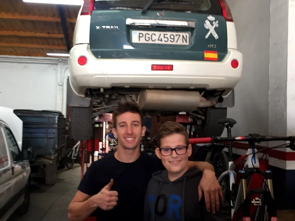 Zwei Personen stehen mit erhobenem Daumen unter einem aufgebockten Auto in der Werkstatt.